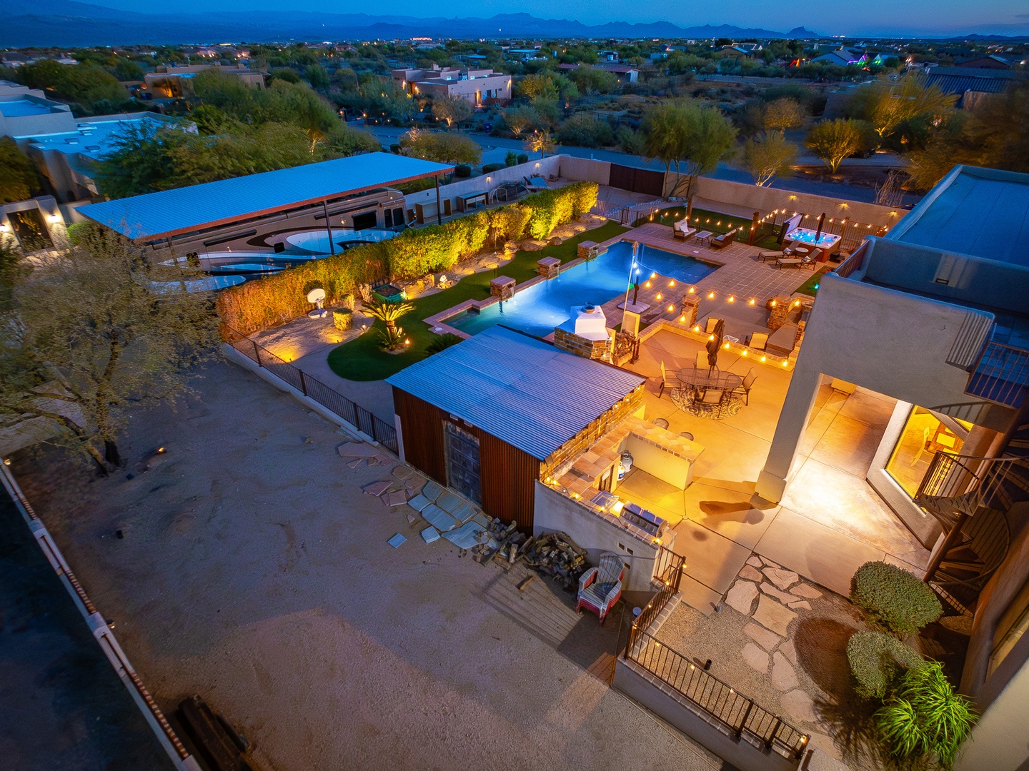 An aerial twilight view showcasing the property's expansive outdoor areas, including a sparkling pool, landscaped grounds, and mountain backdrop in a serene desert neighborhood.