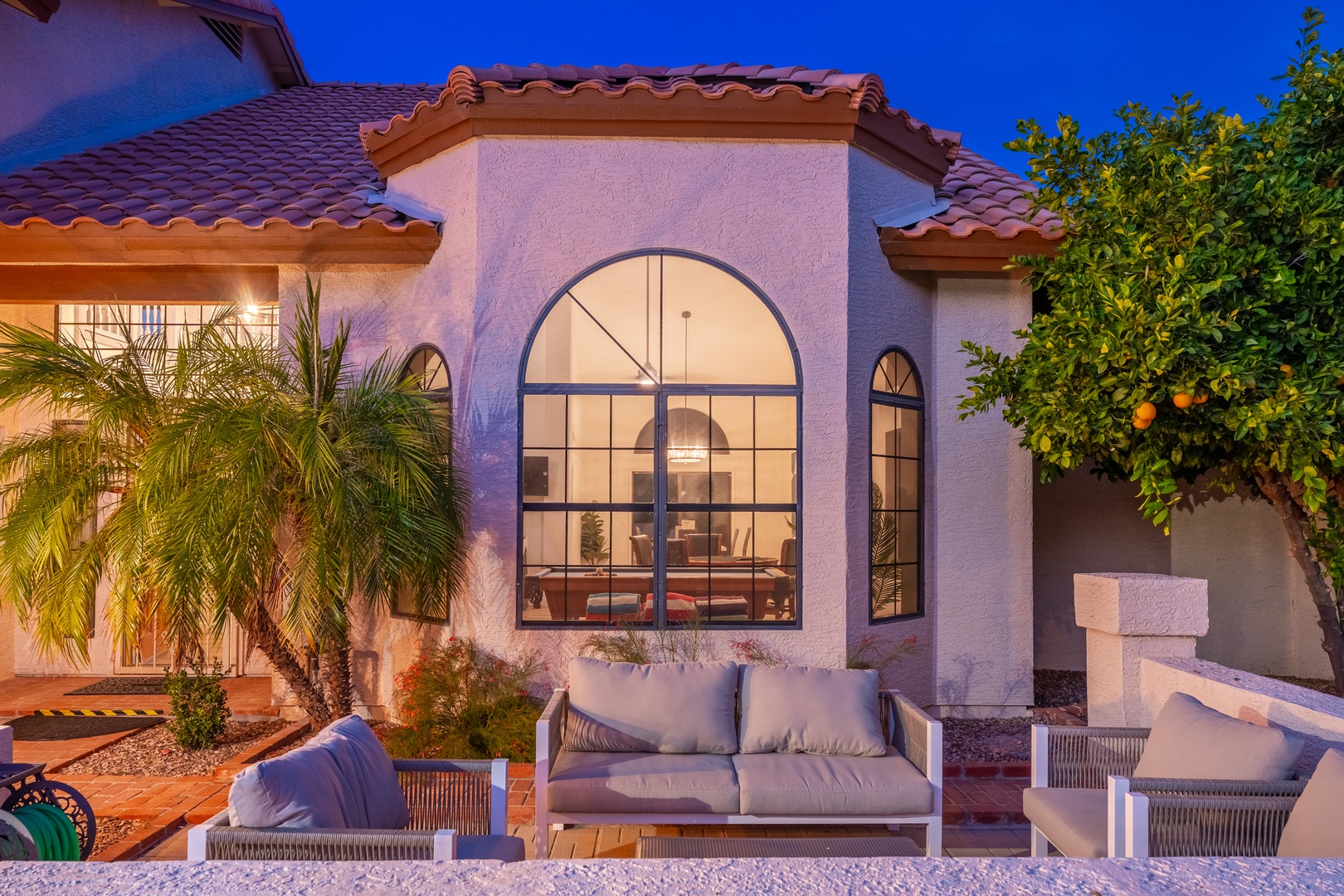 Mediterranean-style villa at twilight features elegant arched windows, terracotta roof tiles, lush tropical landscaping with palms and citrus trees, and inviting patio seating beneath a vibrant blue sky.