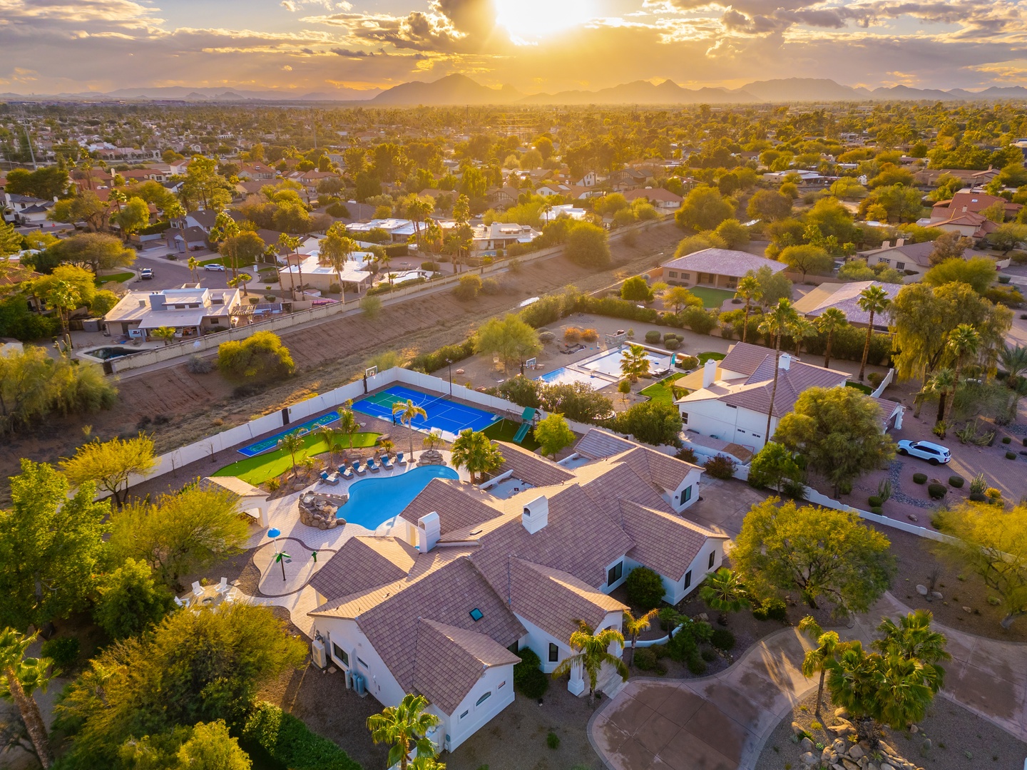 Aerial view of a spacious vacation rental estate at sunset, featuring dual swimming pools and lush landscaping nestled in a tranquil Arizona neighborhood with mountain views.