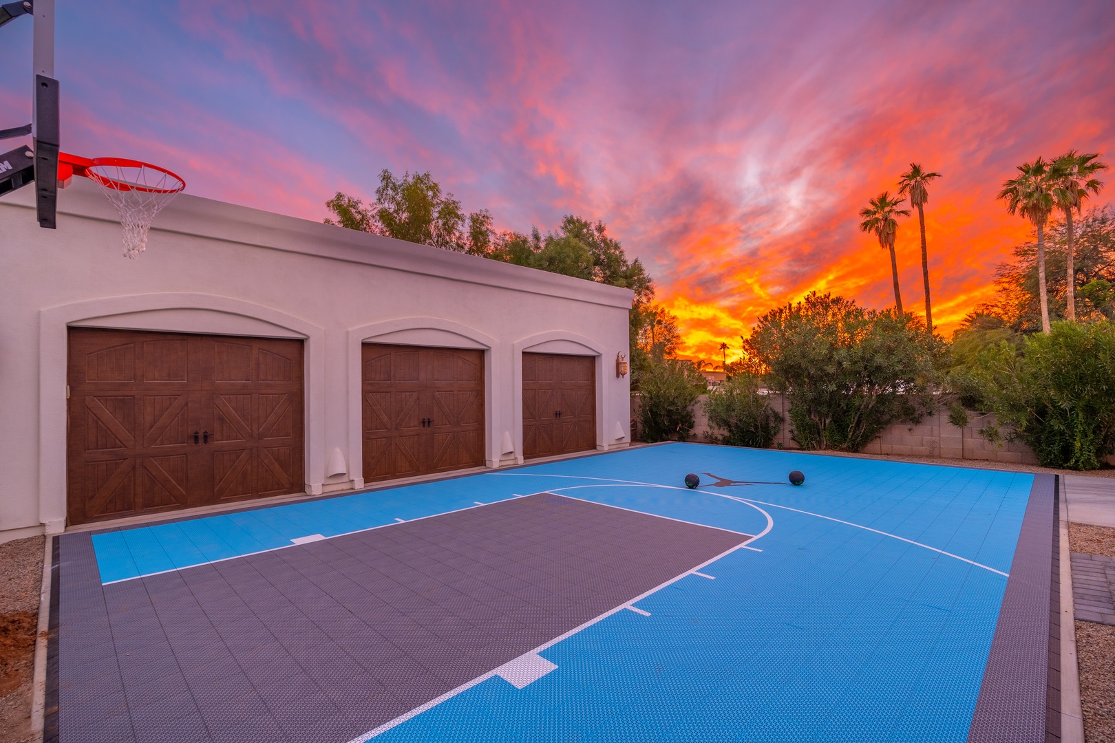 A vibrant sunset paints the sky above the property's private outdoor basketball court, surrounded by palm trees and mature landscaping—an inviting arrival scene in a peaceful desert neighborhood.