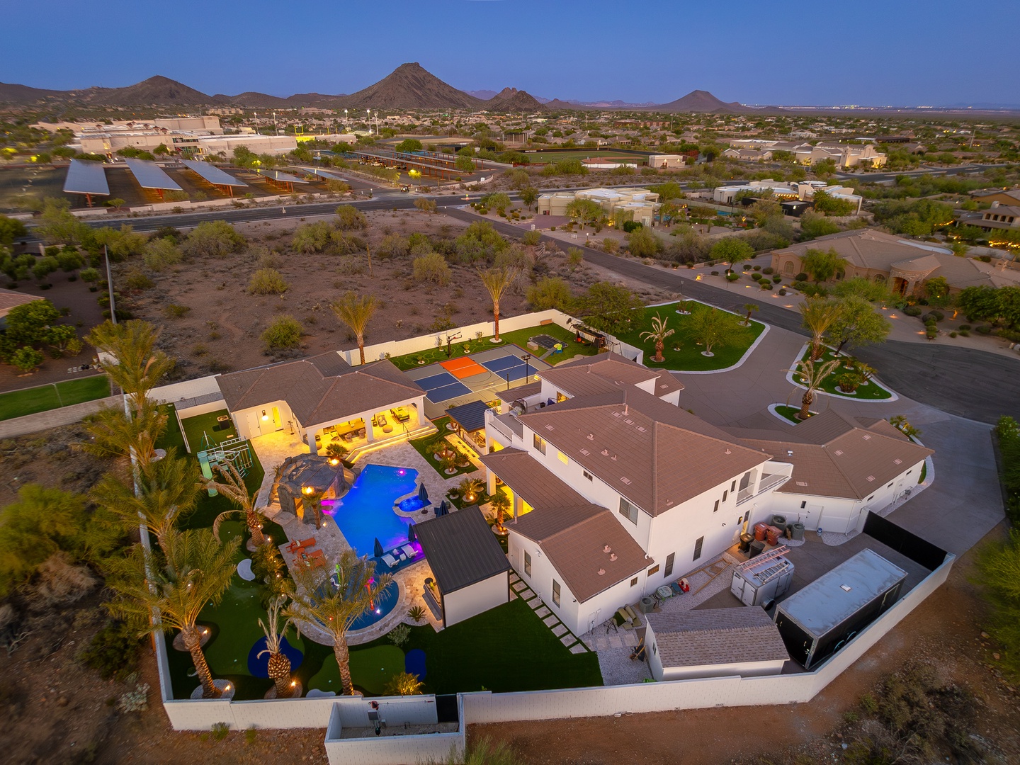 An aerial view captures a luxury desert vacation home nestled in a quiet residential area, featuring a stunning pool, sport court, and expansive outdoor entertainment spaces surrounded by mountain vistas.