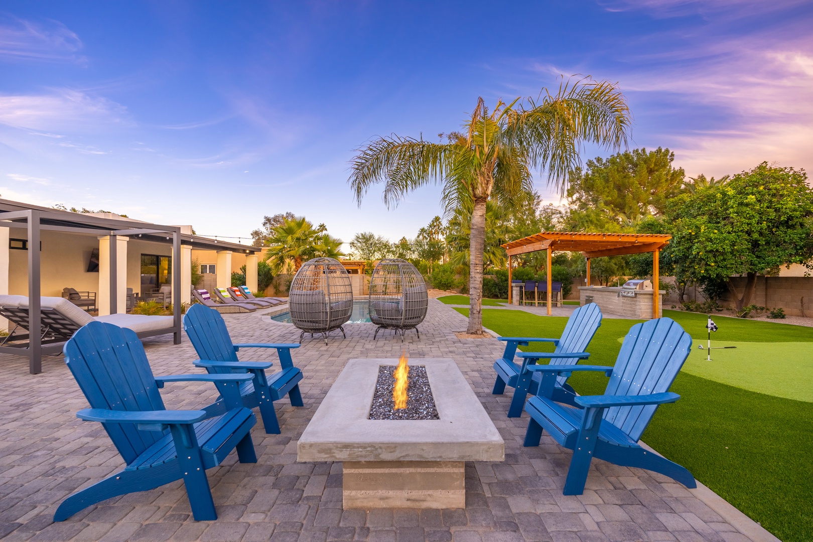 A spacious backyard retreat featuring a modern fire pit surrounded by Adirondack chairs, lush palm trees, putting green, and covered patio—perfect for evening gatherings or quiet relaxation under desert sunsets.
