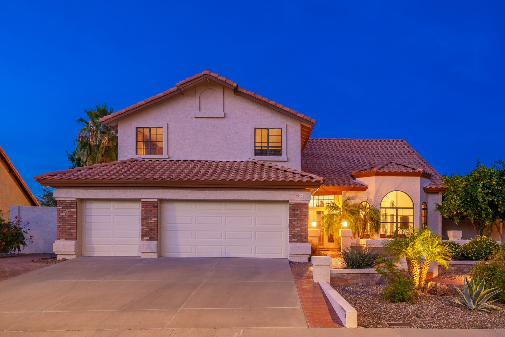 A Southwestern-style property featuring stucco exterior, red tile roof, and desert landscaping with palm trees and agave plants. Warm lighting welcomes arrivals at dusk.