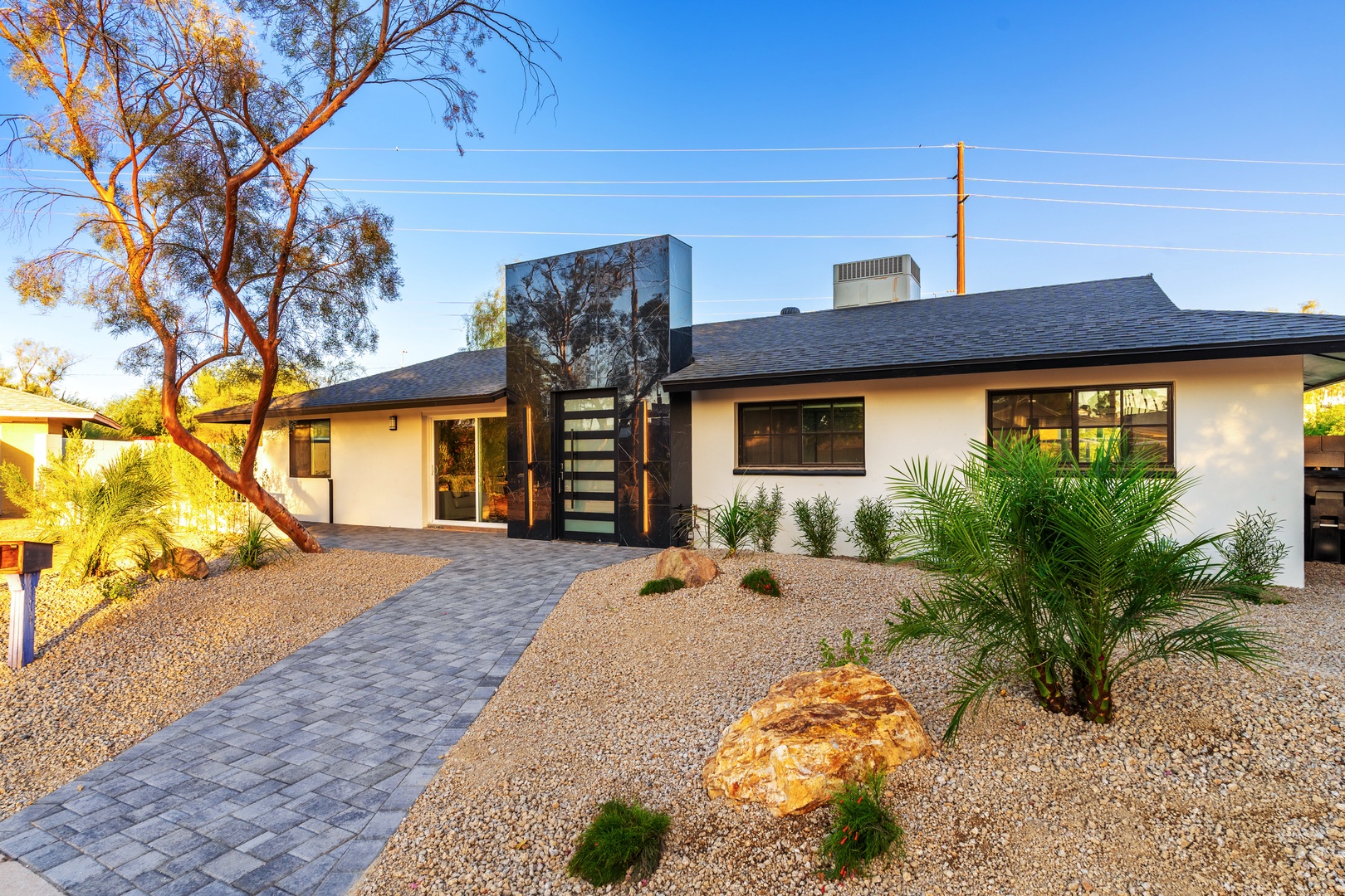 A sleek mid-century modern retreat framed by desert palms and natural stone landscaping. The property's striking entrance gate and paver walkway welcome guests to a beautifully designed desert oasis.