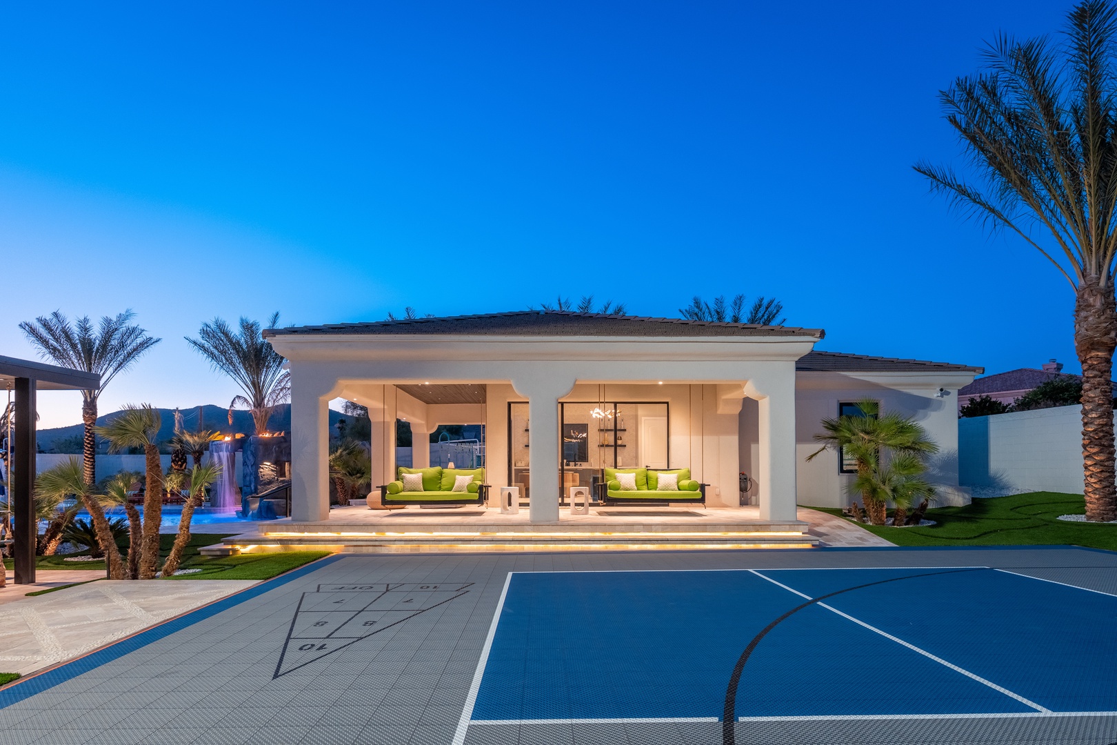 A stunning modern villa features elegant covered outdoor lounges and a sport court, set against desert mountain views and lit palms as twilight descends on this luxury desert oasis.