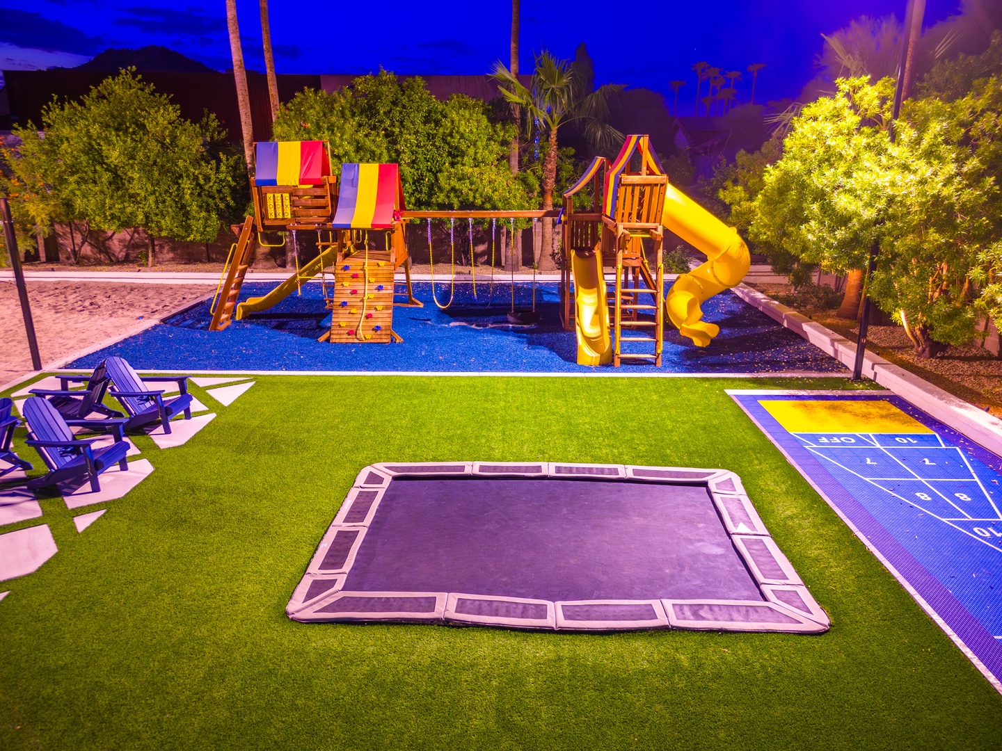 A well-equipped outdoor play area at dusk, featuring a wooden playground with slides and swings, an in-ground trampoline, artificial turf, hopscotch, and lounge seating surrounded by mature landscaping and mountain views.