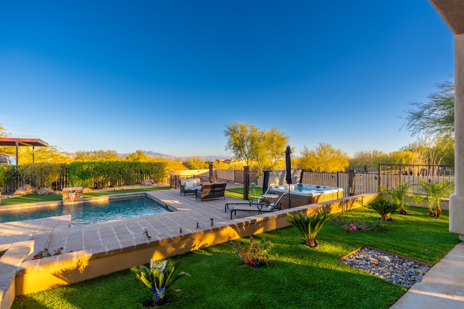 Scenic desert property featuring a swimming pool, hot tub, and landscaped grounds beneath distant mountains and golden-lit trees.
