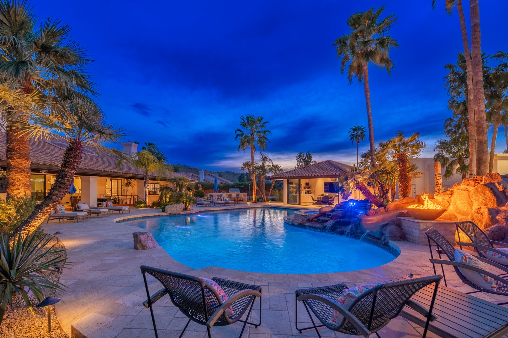 The property's evening ambiance showcases a resort-style pool, tropical landscaping, fire features, and mountain backdrop—a serene desert retreat setting.
