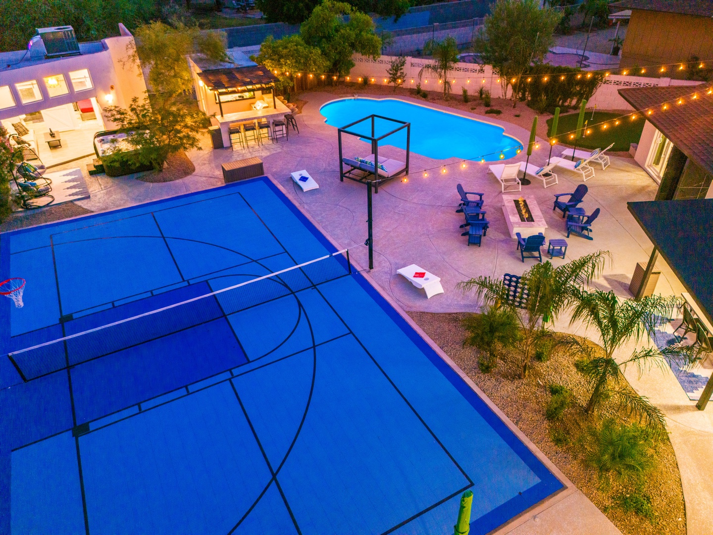 Aerial view of the property featuring a vibrant outdoor recreation area with a full-size basketball court, sparkling pool, and multiple lounge zones under glowing string lights—perfect for active days and relaxing evenings.