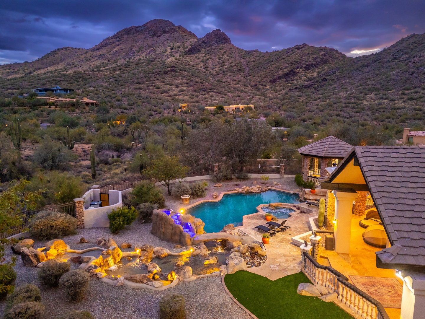 A stunning desert retreat nestled in the foothills features a private pool, spa, and multiple outdoor lounging areas surrounded by mountain views and natural landscape at dusk.