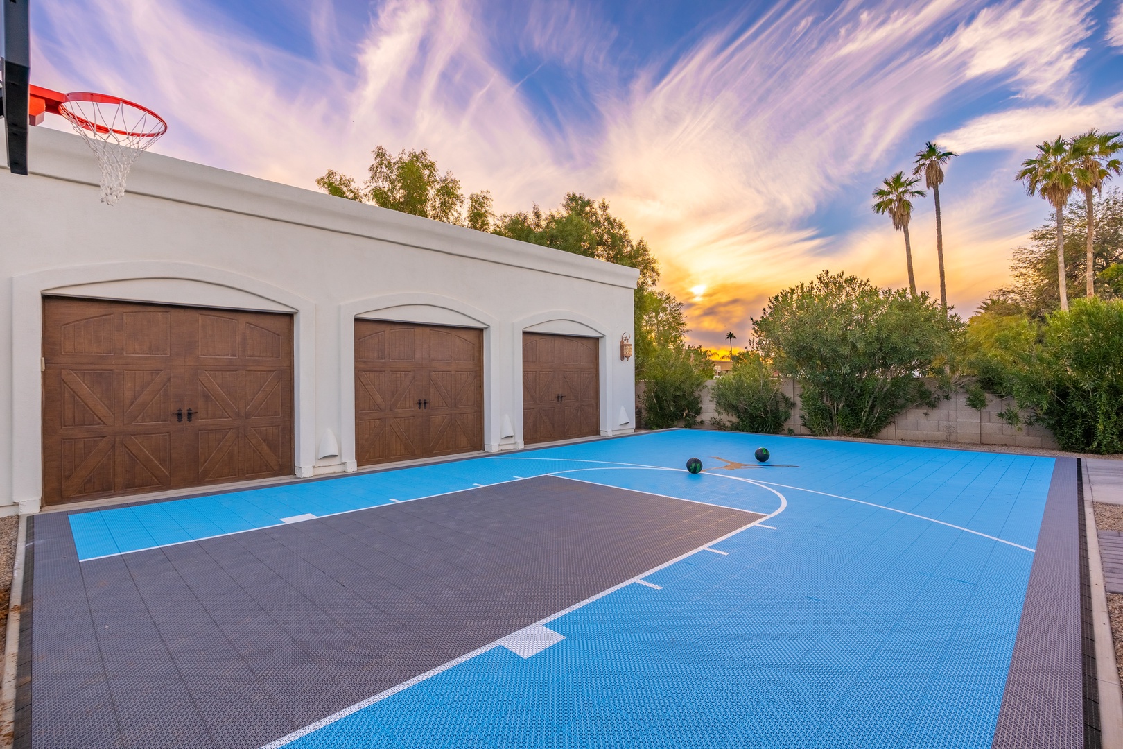 A private half-court basketball area with hoop and fencing, backdropped by palm trees and a stunning sunset over the desert landscape.