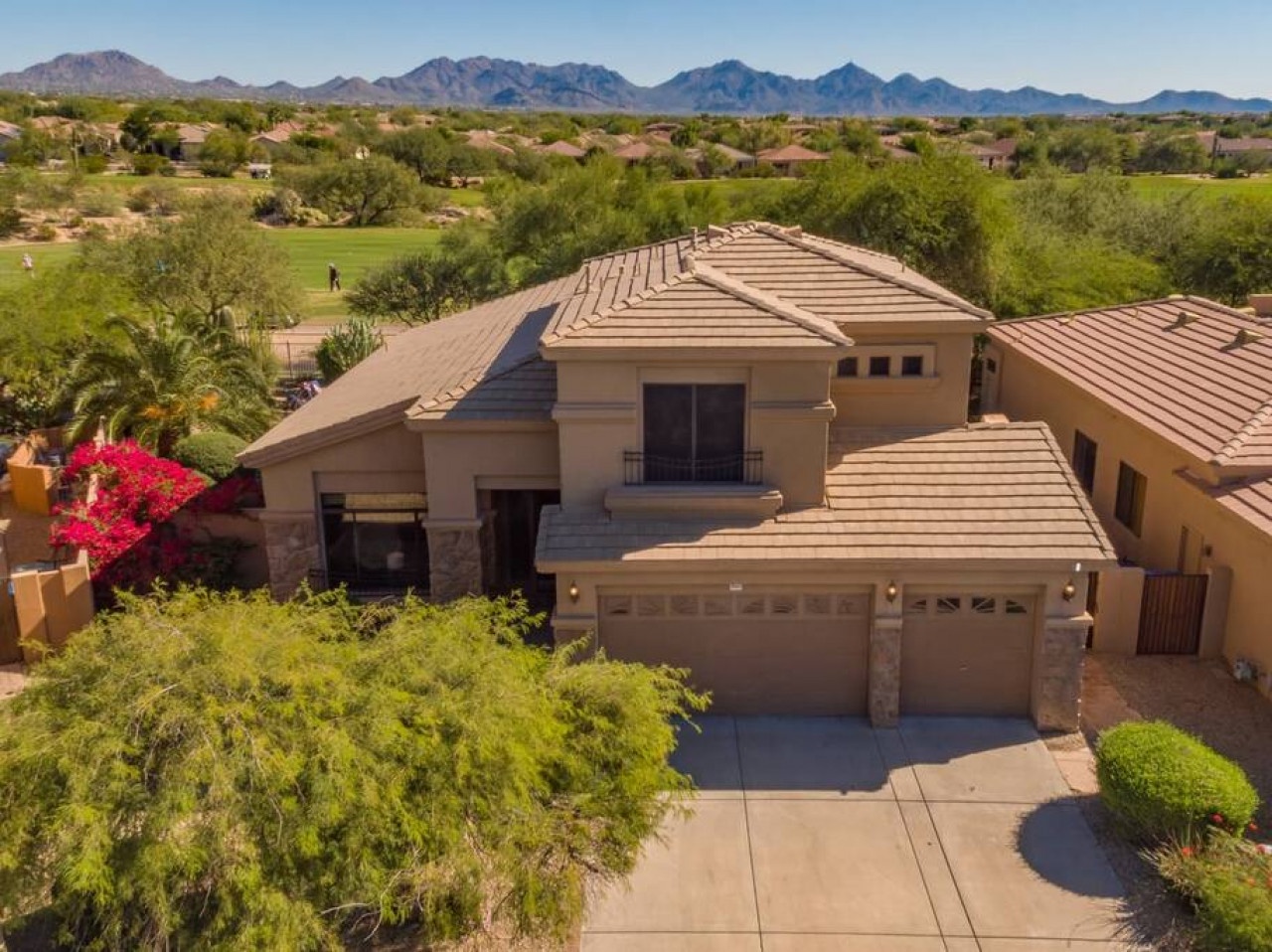 Modern desert home with tile roof sits amid lush landscaping, overlooking a golf course with mountain views beyond.