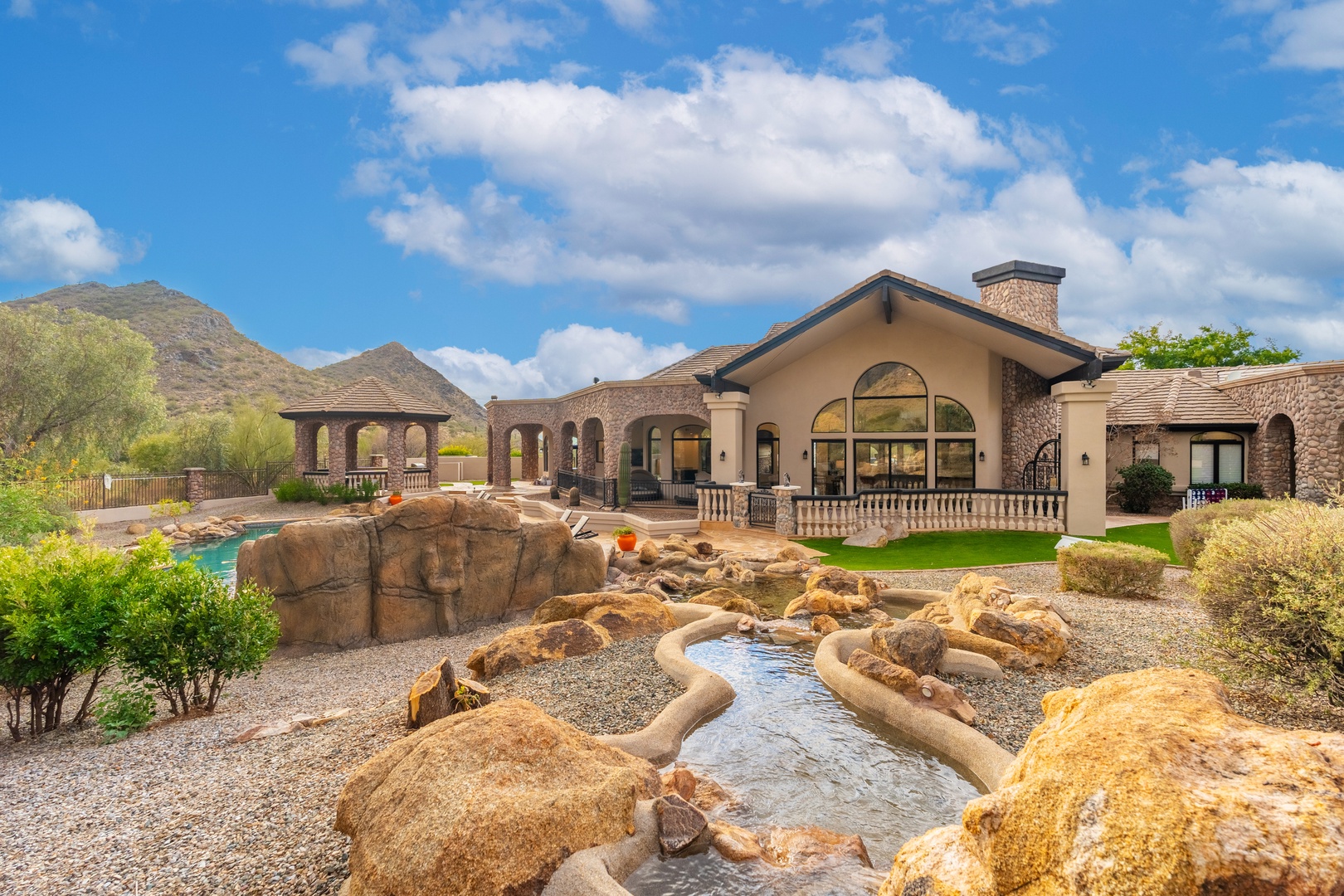 Stunning desert estate featuring natural rock water features, covered outdoor pavilions, and manicured landscaping framed by mountain vistas. An oasis of luxury and tranquility awaits.