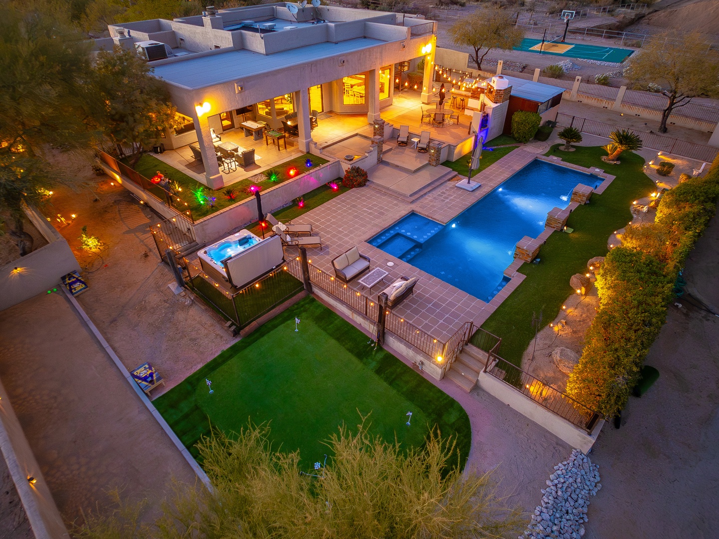 Stunning contemporary desert retreat featuring expansive outdoor living spaces, resort-style pool with hot tub, multiple dining and lounging patios, and beautifully landscaped grounds illuminated at twilight.