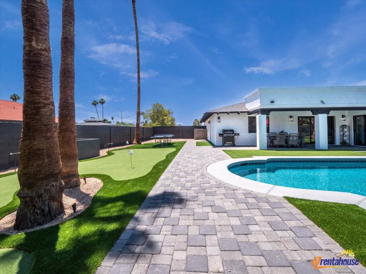 Private desert oasis with sparkling pool, manicured lawn, and covered patio area surrounded by towering palms under endless blue skies.