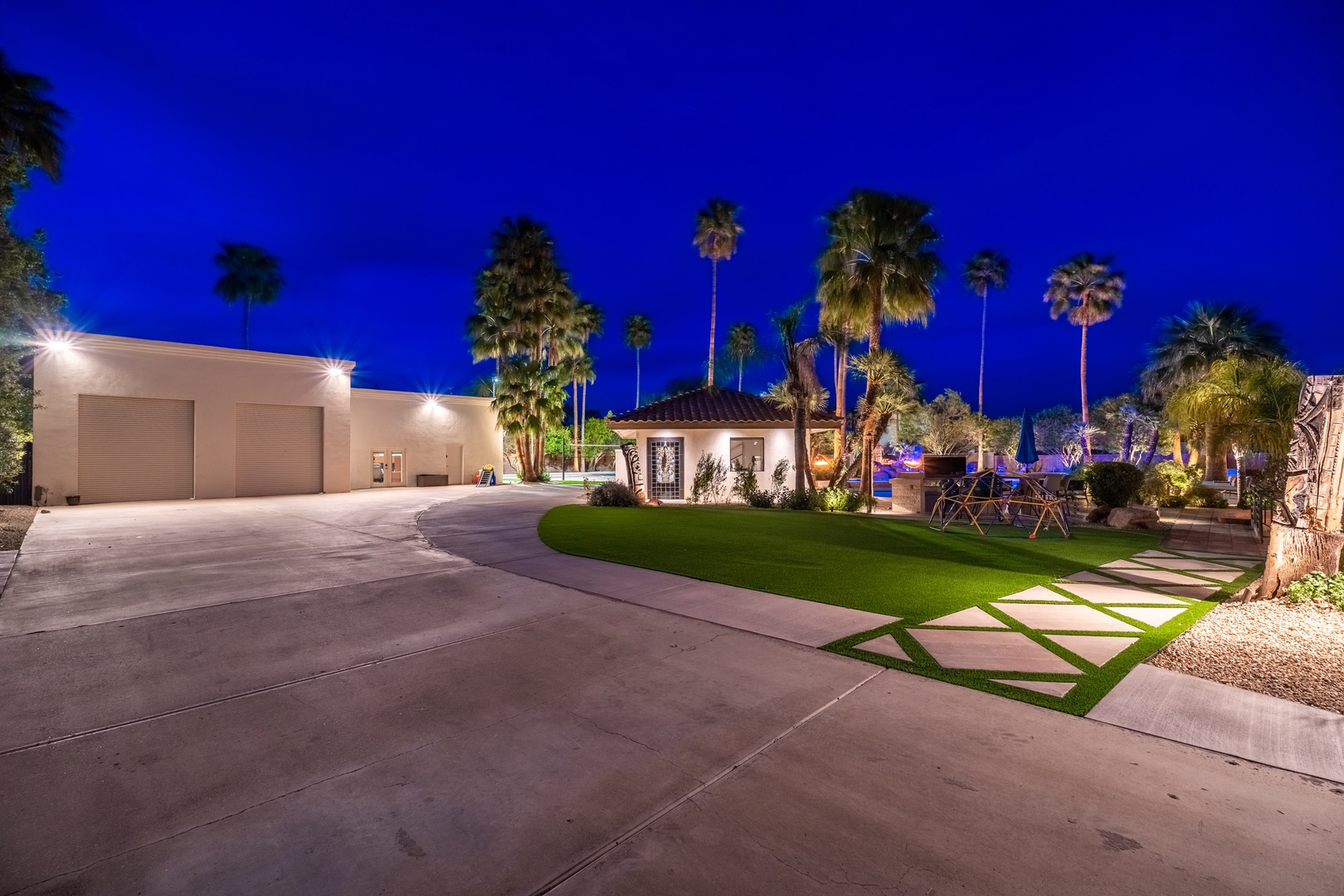 An elegantly lit property exterior featuring manicured lawns, palm trees, and modern architecture set against a twilight sky. This desert oasis offers ample parking and stylish outdoor spaces.