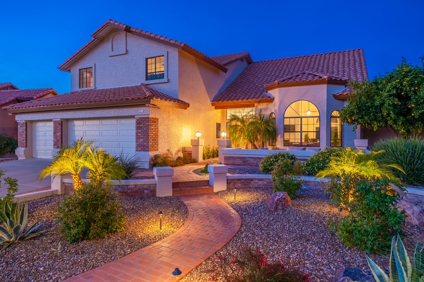 A beautifully lit two-story vacation home with Spanish tile roofing and landscaped walkways welcomes guests at twilight, showcasing warm exterior lighting and desert-style gardens.