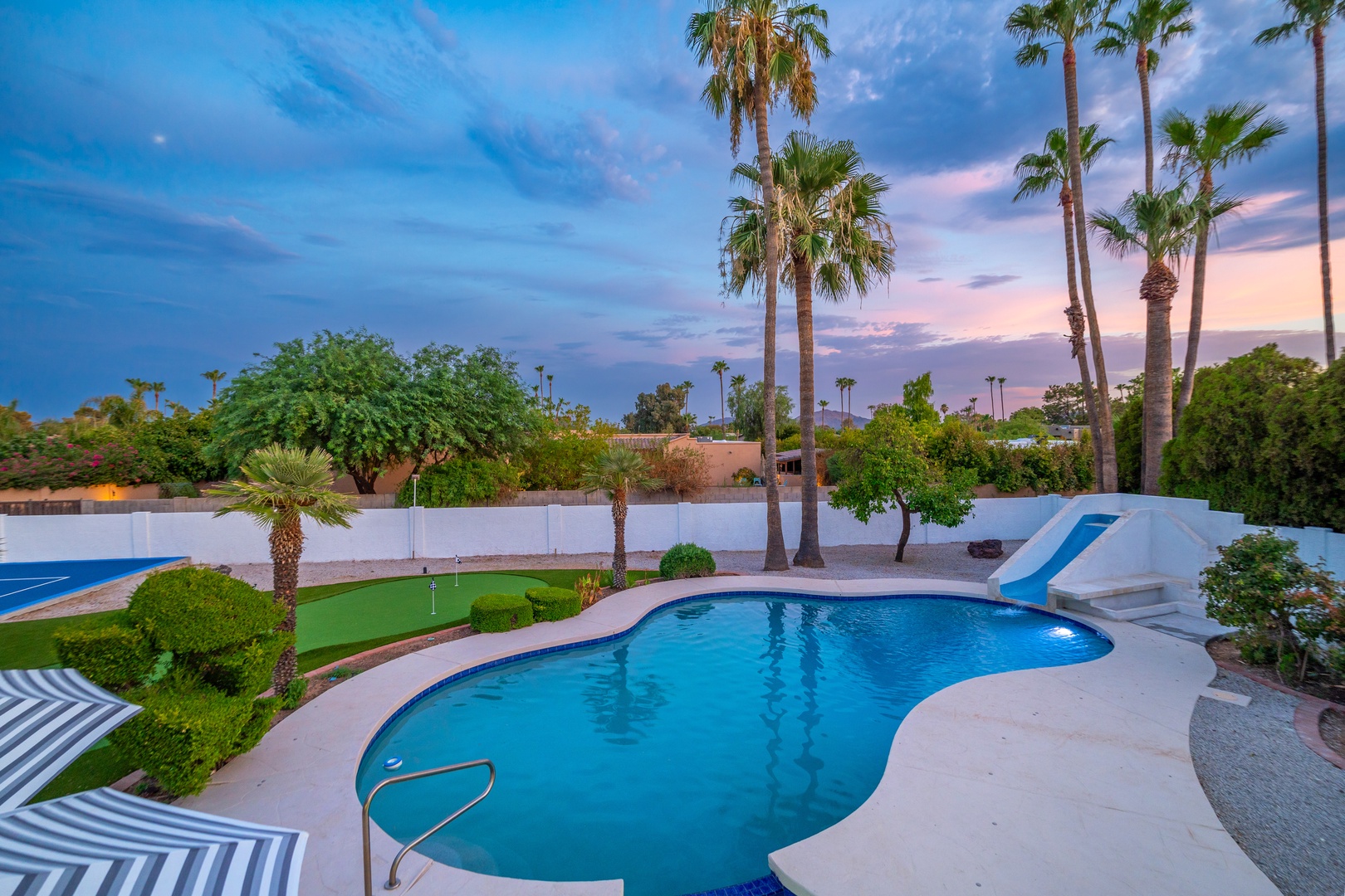 Spacious backyard oasis features a sparkling pool, putting green, and towering palms set against a stunning twilight sky. The perfect retreat for desert relaxation.