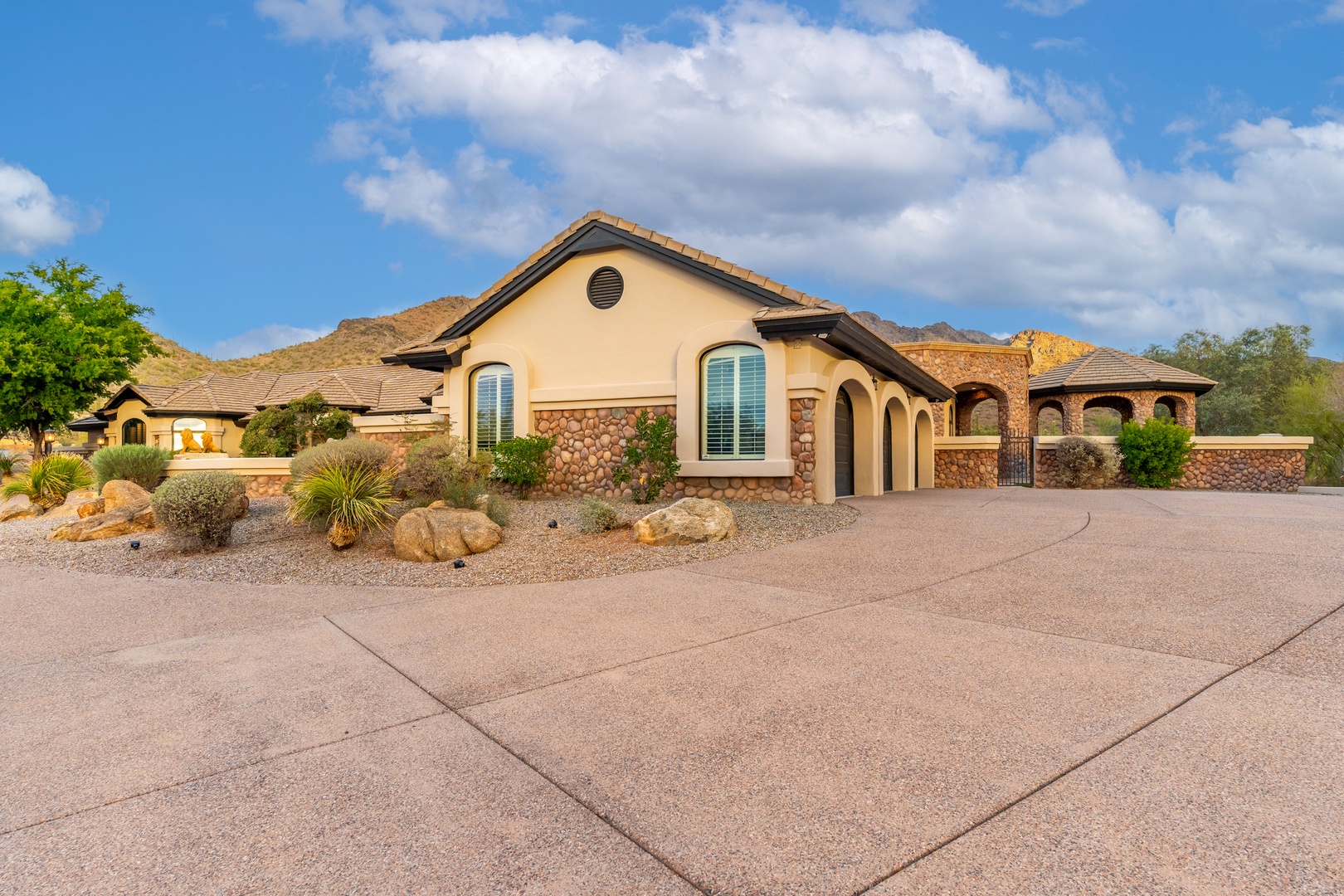 This Mediterranean-style property features warm stucco exteriors, stonework accents, and desert landscaping with native plants and large boulders, creating an inviting arrival experience in a peaceful neighborhood setting.