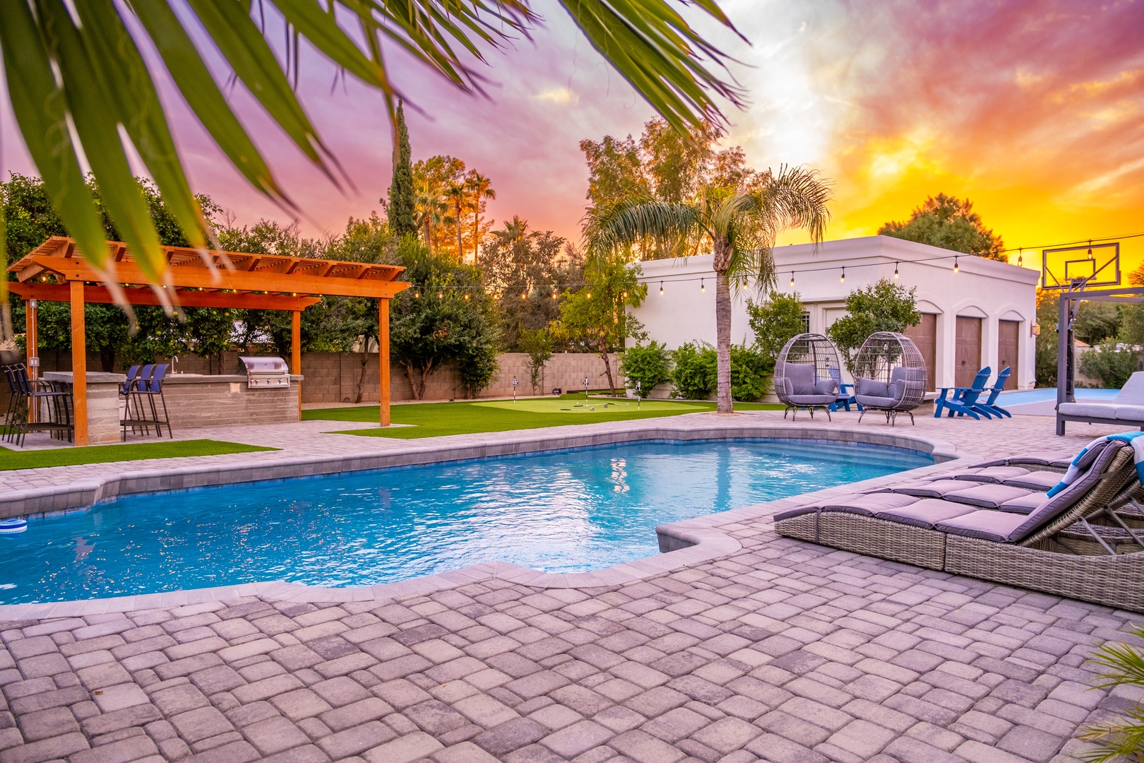 A stunning Arizona sunset bathes the resort-style backyard in golden light, showcasing a sparkling pool, cozy loungers, and a shaded barbecue area—perfect for unwinding with friends and family.