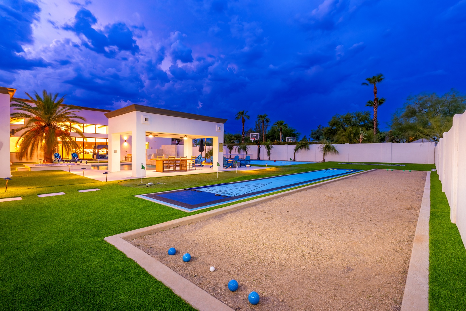 A stunning backyard retreat with a private bocce court, pool with basketball hoop, and covered patio awaits your perfect vacation evening beneath dramatic twilight skies.