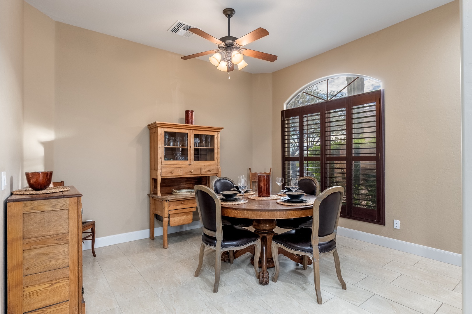 Gather around this elegant round table for memorable meals bathed in natural light from the arched window. Warm wood furnishings and a charming hutch create an inviting space perfect for your morning coffee or evening dinner.