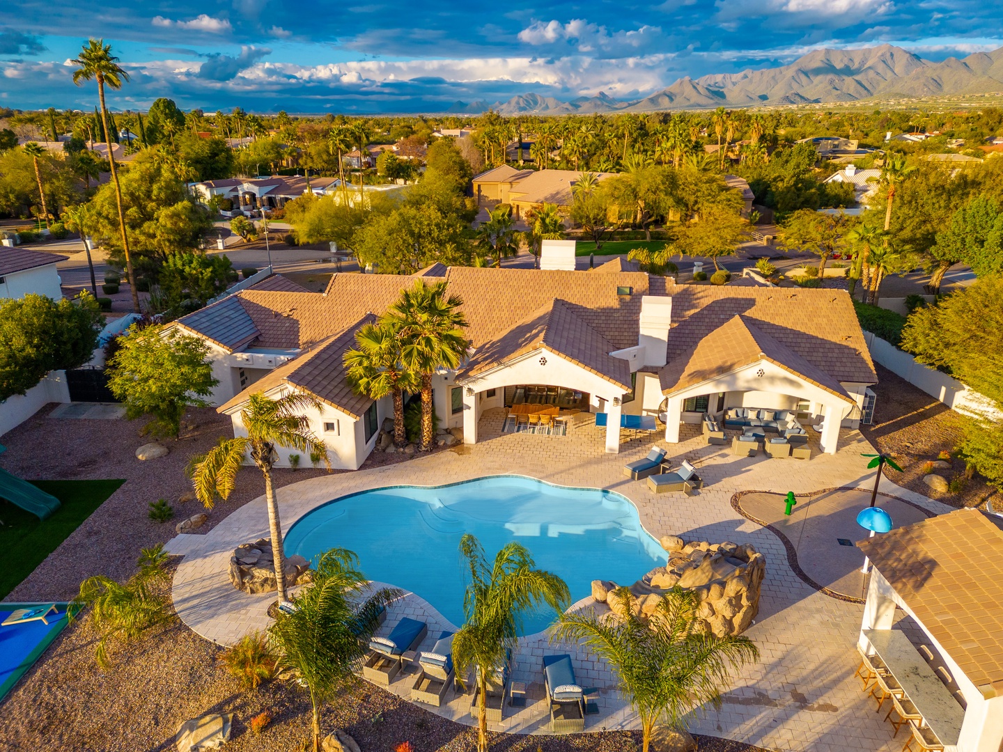 Aerial view of the vacation rental property featuring a sparkling pool, covered patio, and panoramic mountain vistas in a quiet desert neighborhood with palm trees and clear Arizona skies.