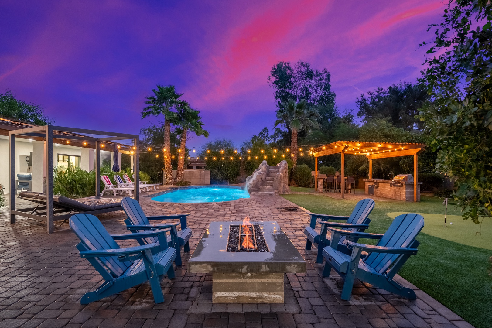 A stunning dusk view reveals the property's expansive outdoor living spaces with pool, fire pit, and pergola-covered dining area set against a vibrant desert sky.