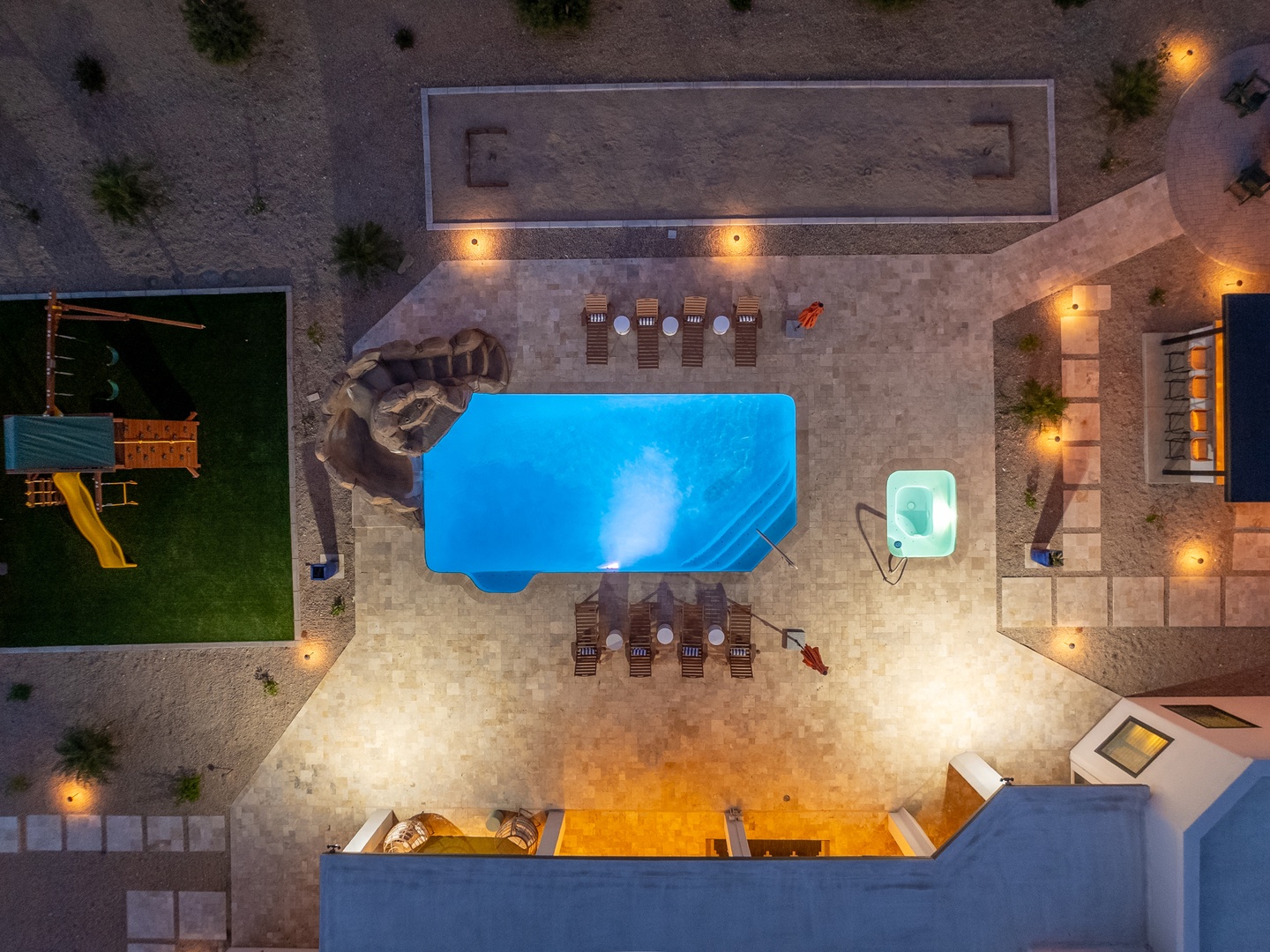 Aerial view showcasing the property's outdoor amenities at dusk, featuring a sparkling pool, surrounding patio with lounge chairs, small hot tub, children's play area, and beautifully illuminated grounds with Mediterranean-style landscaping.
