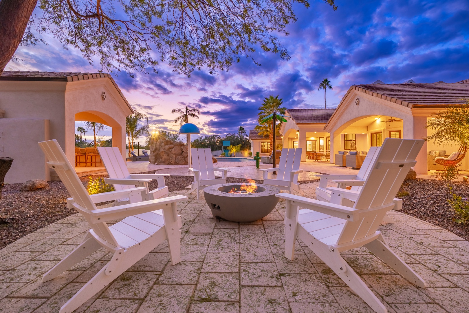 Stunning twilight patio featuring a circular fire pit surrounded by white Adirondack chairs, with palm trees framing a desert resort-style courtyard, illuminated casitas, and dramatic painted skies overhead.