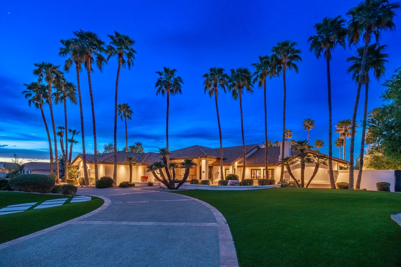 A luxury desert estate framed by towering palms, illuminated against a dramatic twilight sky with manicured grounds and a grand entrance driveway welcoming arrivals.