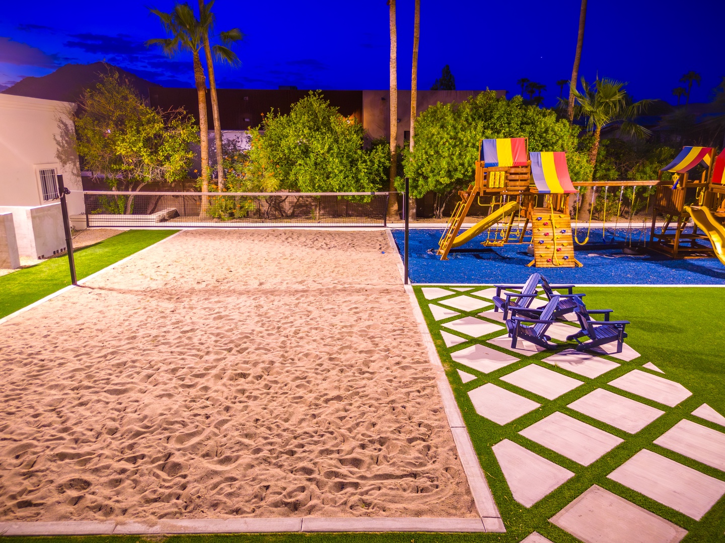 This private backyard features a sand volleyball court, colorful playground, and seating area under palm trees—perfect for family fun and evening gatherings beneath the desert sky.