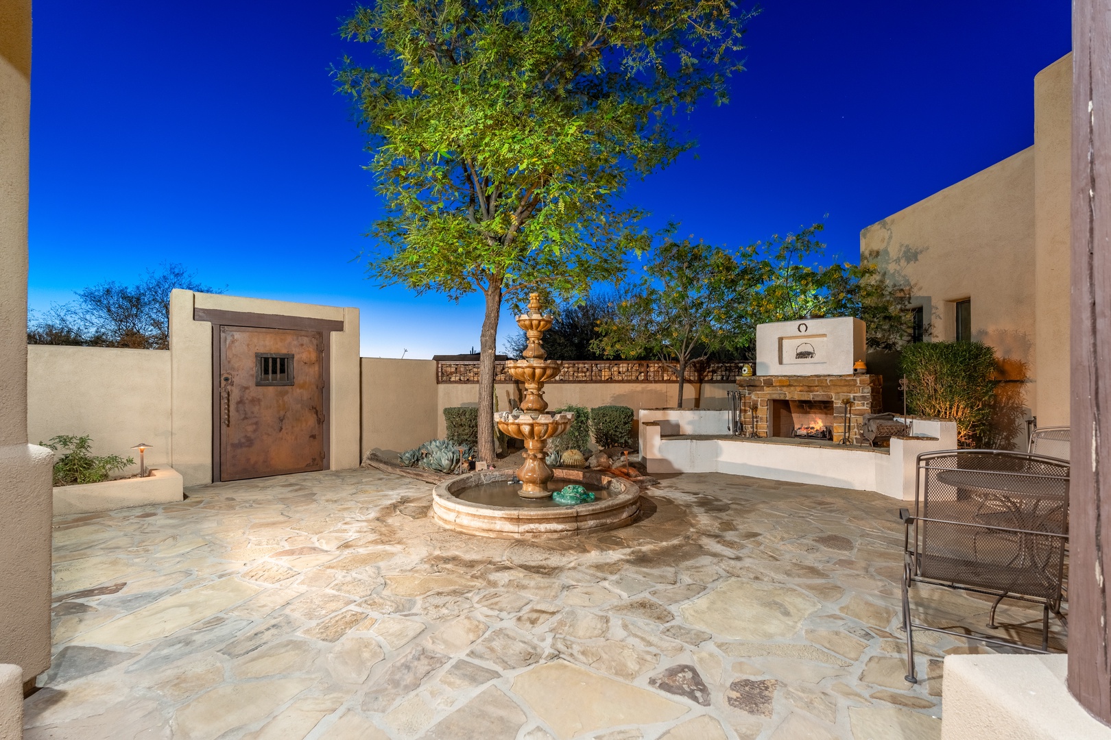 A serene courtyard features an ornate tiered fountain, flagstone paving, and a cozy outdoor fireplace, all framed by desert landscaping beneath a brilliant blue sky.