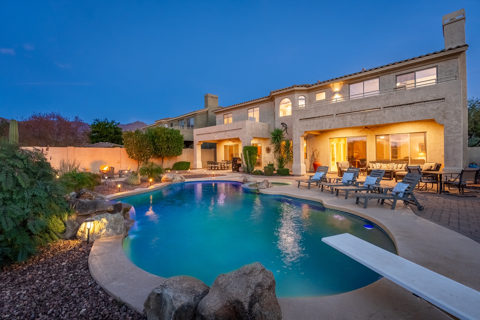 Stunning two-story desert retreat with an inviting illuminated pool, lush landscaping, and multiple outdoor lounging areas set against mountain views at twilight.