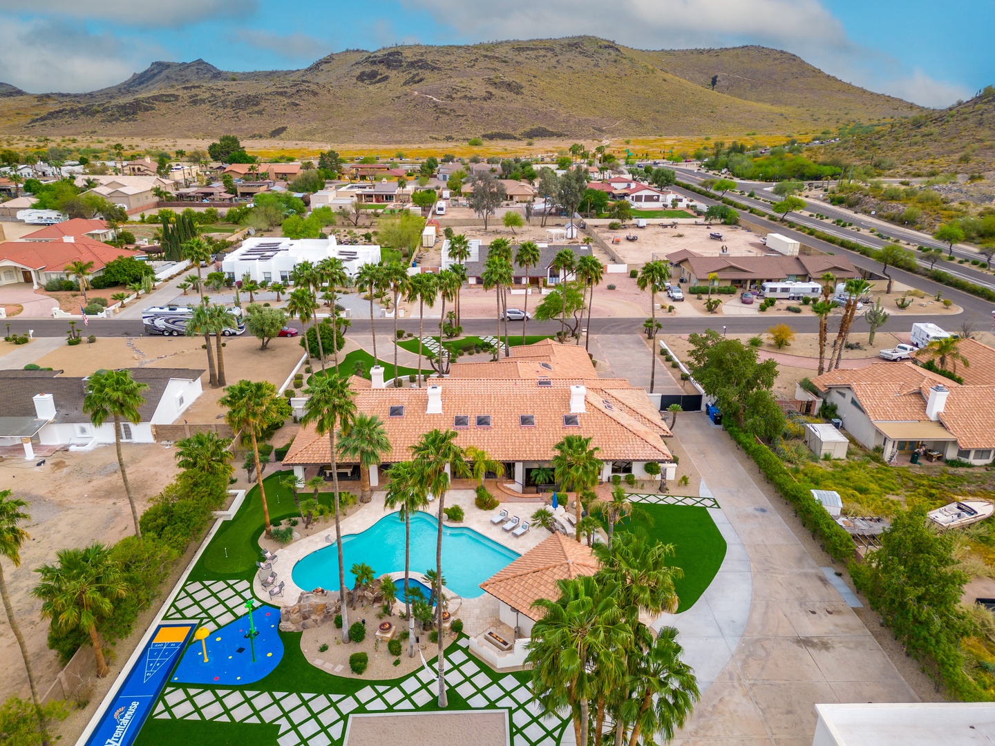 Aerial view of a resort-style property nestled in a desert landscape, featuring a sparkling pool, lush palm trees, and mountain vistas framing the neighborhood.
