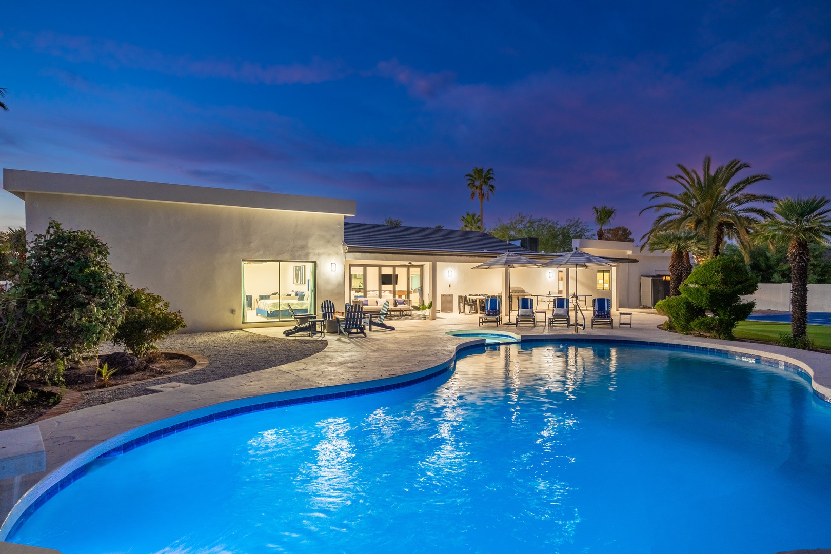 Modern desert retreat with sparkling pool, illuminated at twilight, framed by swaying palms and mountain views in the surrounding area.