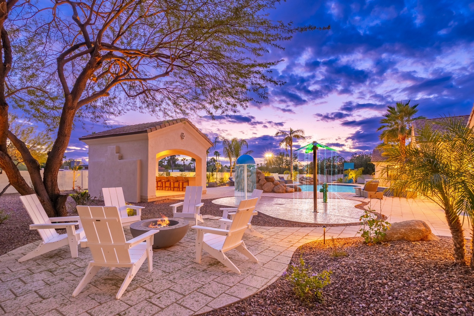 A desert oasis backyard at twilight featuring a pool, splash pad, fire pit, and covered bar area surrounded by palms and mountain views.