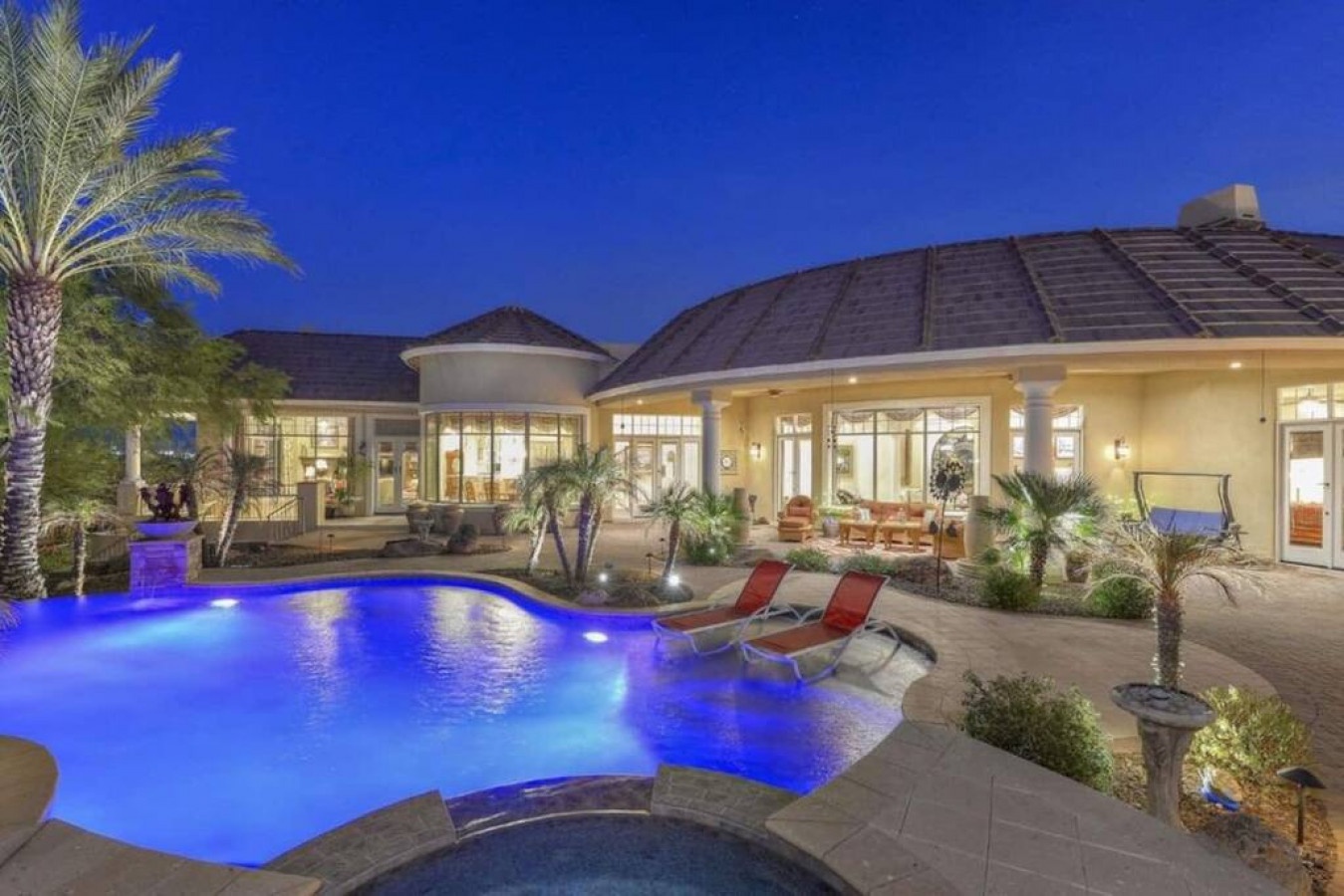 Luxurious evening retreat featuring a beautifully lit pool with vibrant blue lighting, tropical landscaping, and spacious patio areas perfect for relaxing under the stars.