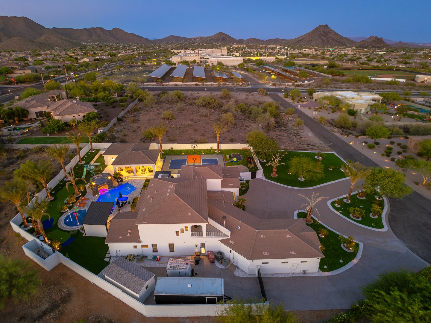 Aerial view of a spacious desert property featuring multiple pools, tennis court, and lush landscaping set against dramatic mountain scenery at dusk.