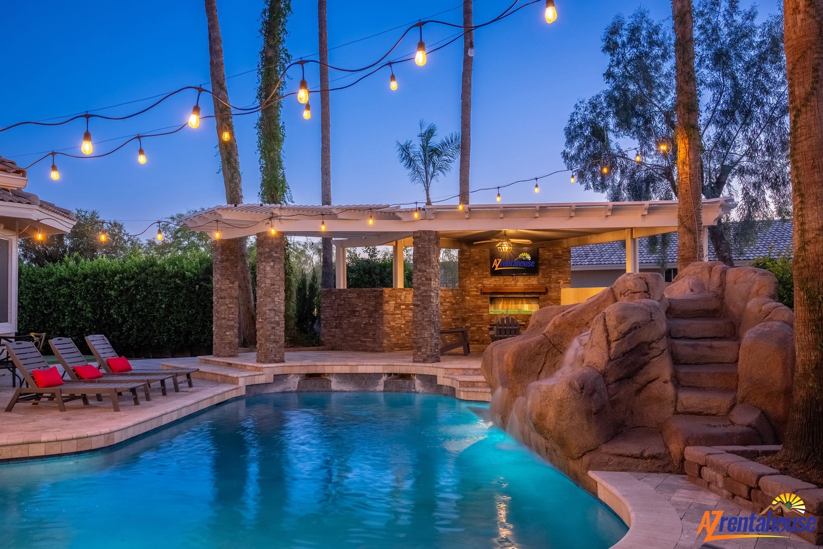 A resort-style backyard oasis featuring a sparkling pool with custom rock waterfall, outdoor kitchen with pergola, string lights overhead, and lounge chairs set against lush desert palms.