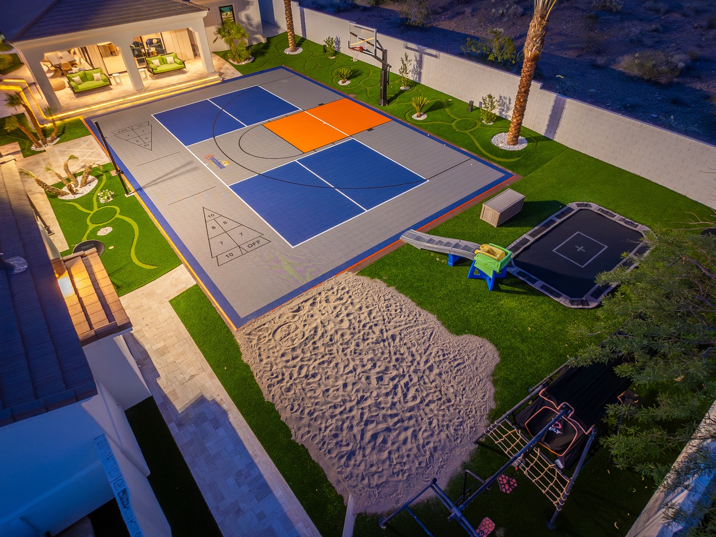 Aerial view showcasing the property's extensive recreational amenities including a regulation basketball court, sand play area, trampoline, and beautifully landscaped grounds with palm trees adjacent to waterfront.