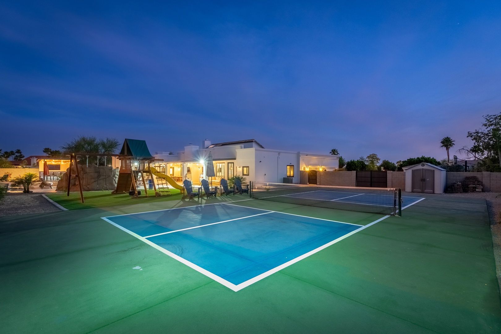 Twilight setting showcasing the private tennis court, playground, and illuminated modern home—ideal for active family getaways and evening outdoor enjoyment.