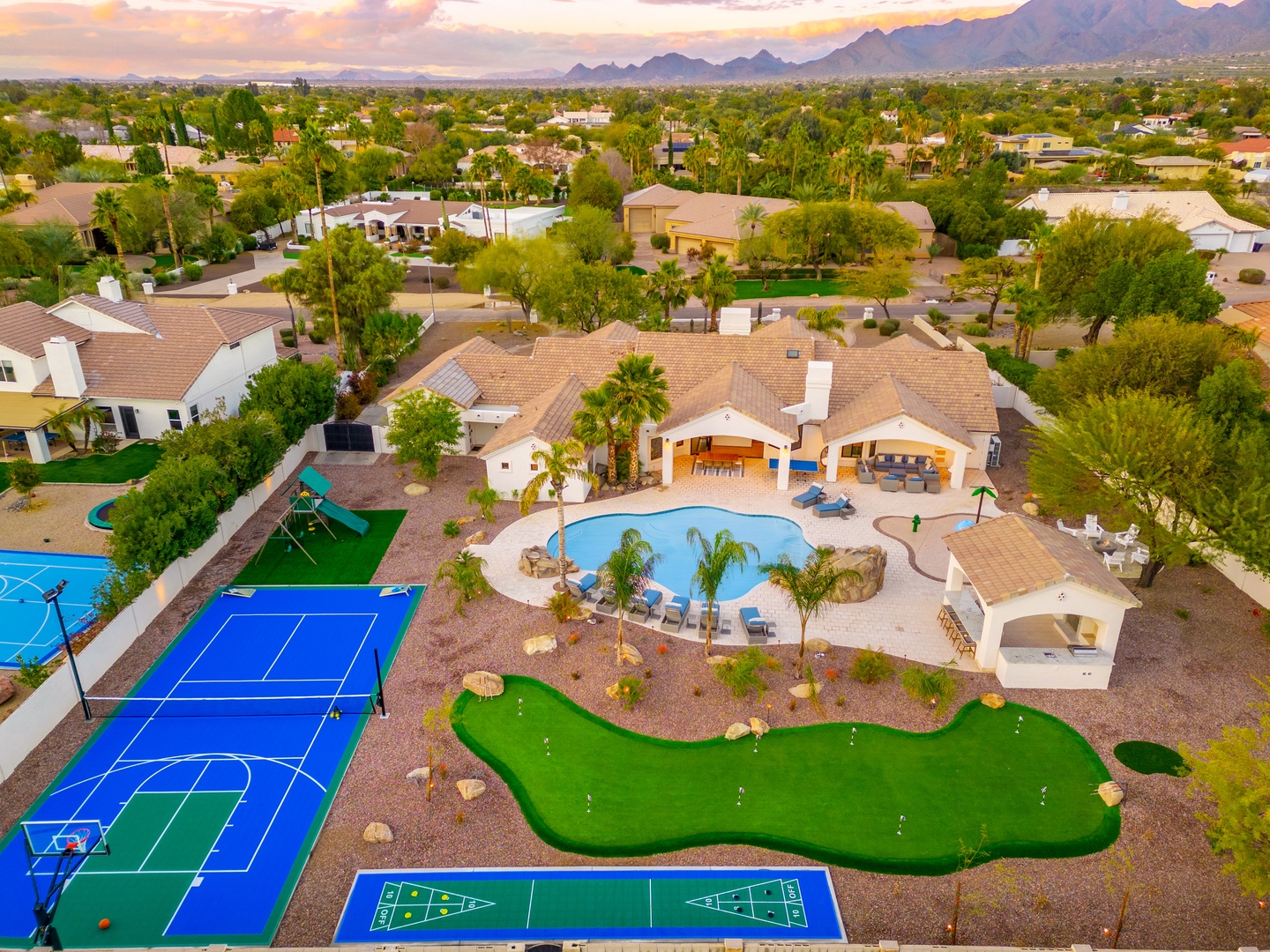 Aerial view of a resort-style property featuring a pool, mini golf, tennis and sport courts, nestled in a lush desert neighborhood with mountain views at sunset.