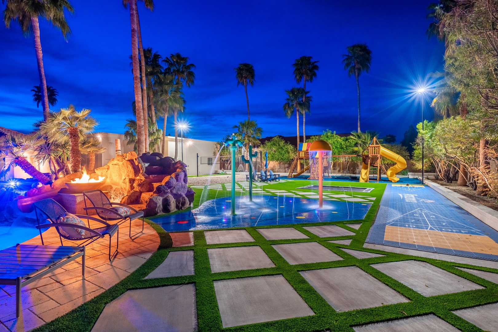 The backyard features a splash pad, playground with slide, fire pit seating, and towering palm trees under evening lights—an entertainer's dream setting.