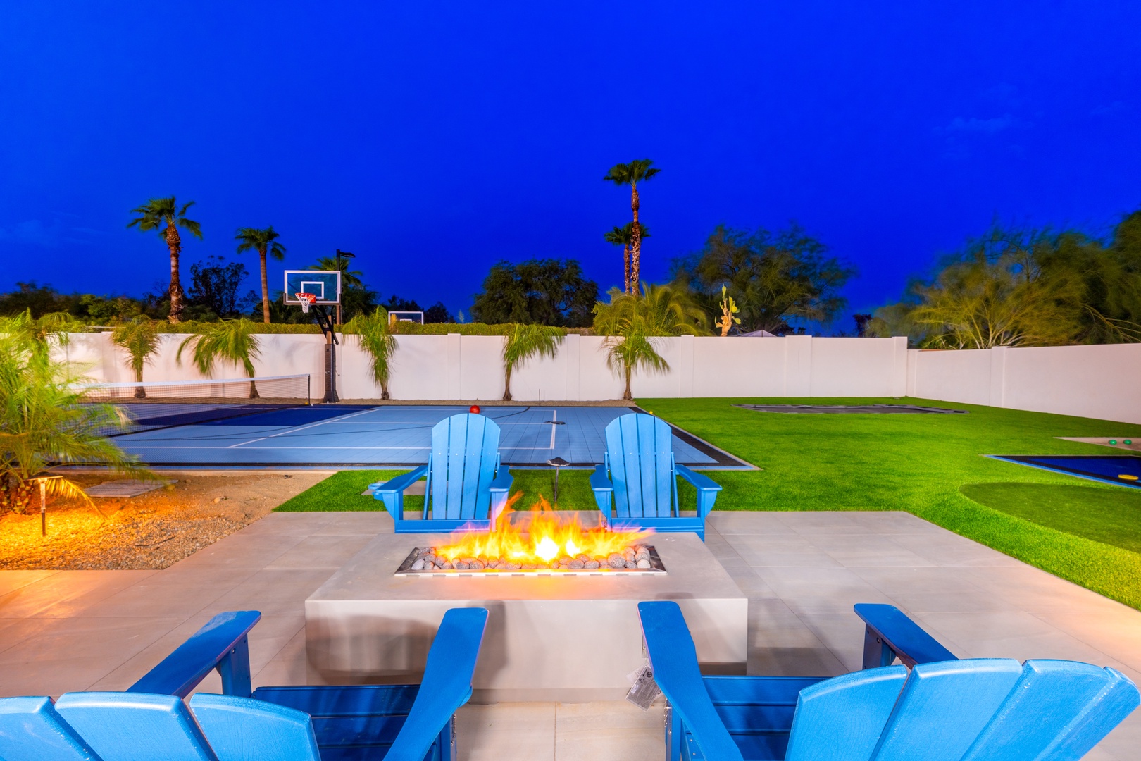 This modern outdoor living space features a striking fire pit surrounded by lounge chairs overlooking the pool, basketball court, and lush landscaping—all under dramatic evening skies.