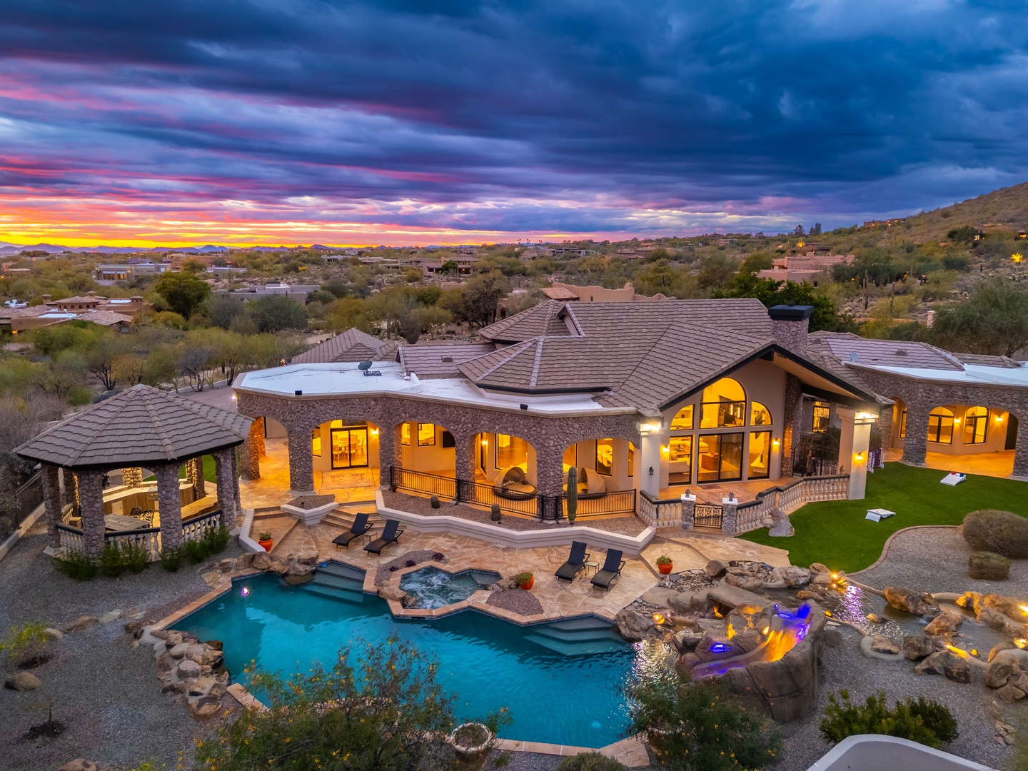 A stunning desert estate at twilight, featuring a custom pool, spa, covered gazebo, and lush lawns set against dramatic Sonoran mountain vistas and wide-open skies.