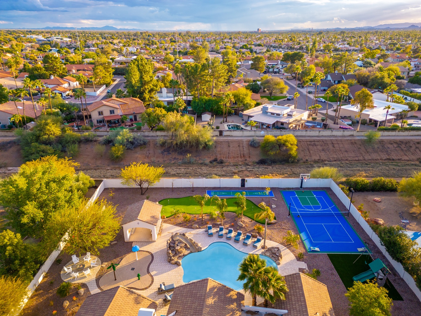 Aerial view of the property showcasing an expansive outdoor oasis with a sparkling pool, sports courts, and entertainment areas set against a backdrop of the surrounding residential neighborhood and distant mountain ranges.