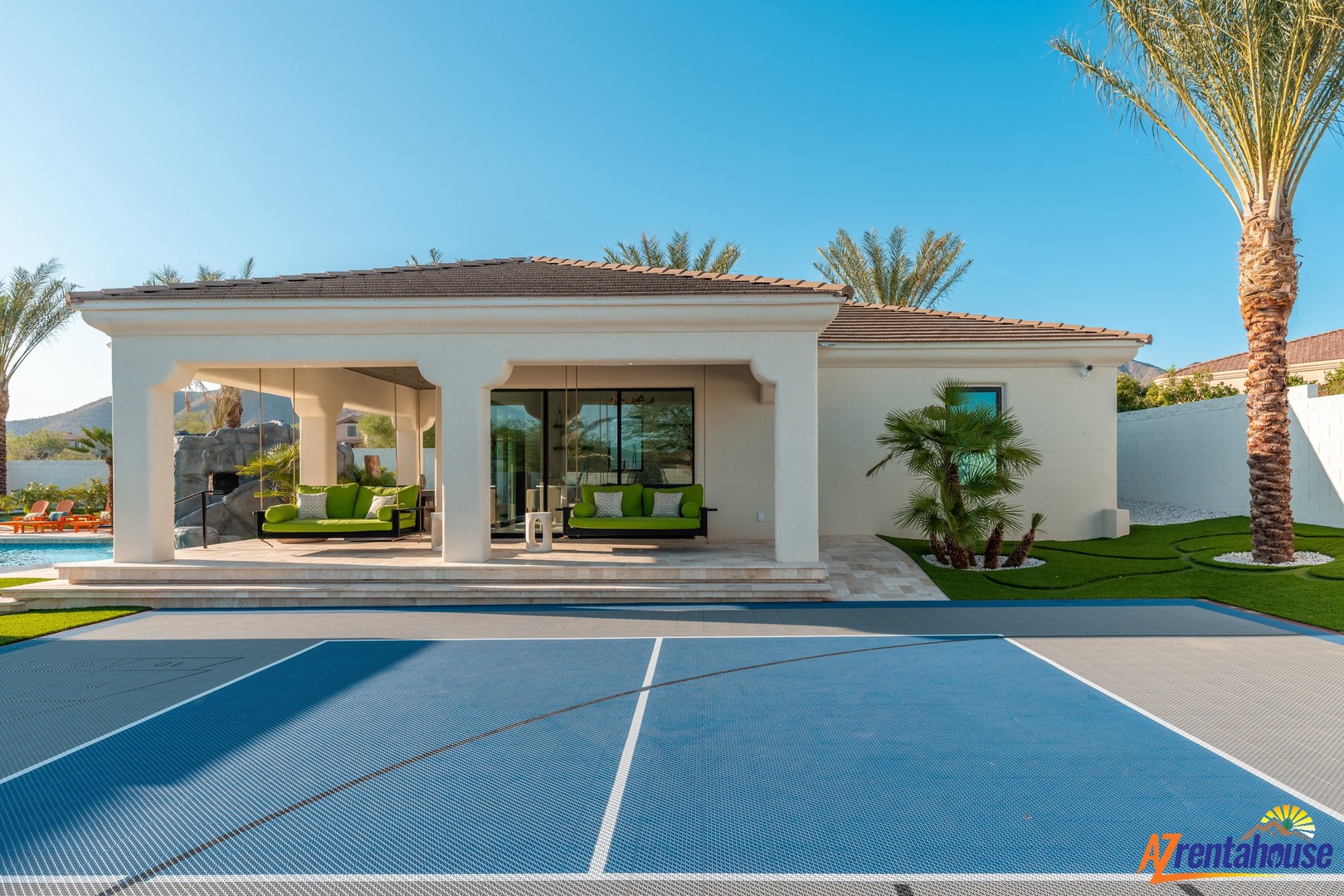 Modern white villa with blue tennis court, covered poolside terrace, and tropical palm trees in a sunny resort setting.