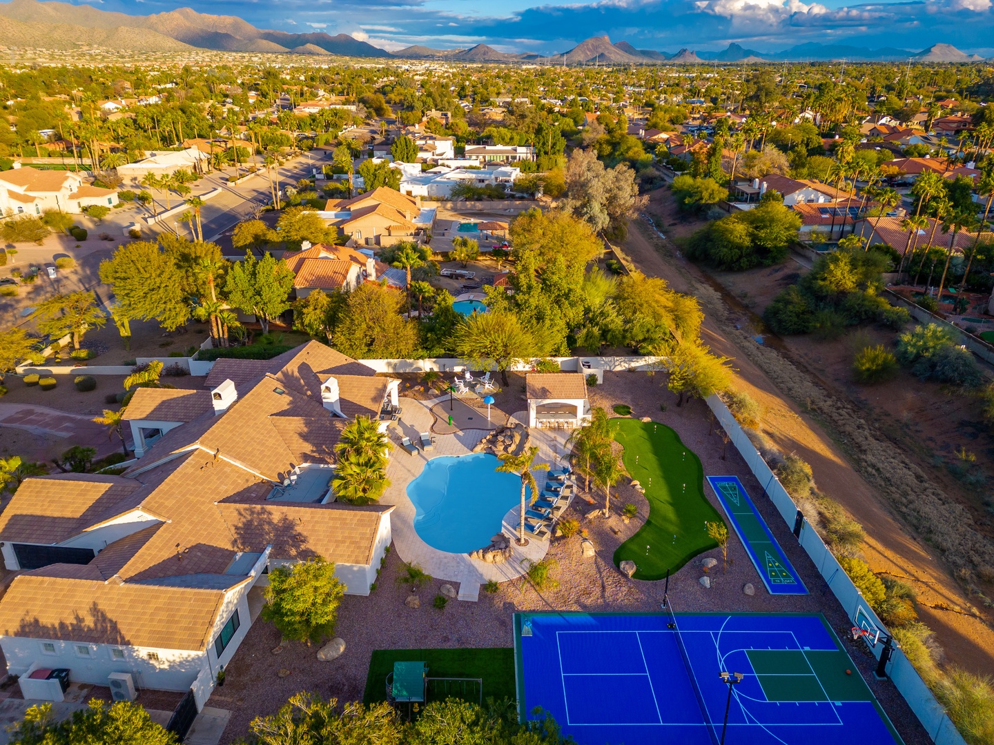 Expansive estate featuring a sparkling pool, vibrant tennis and pickleball courts, and lush landscaping, set against a mountain-framed desert neighborhood. A private resort-style property offering abundant outdoor recreation.