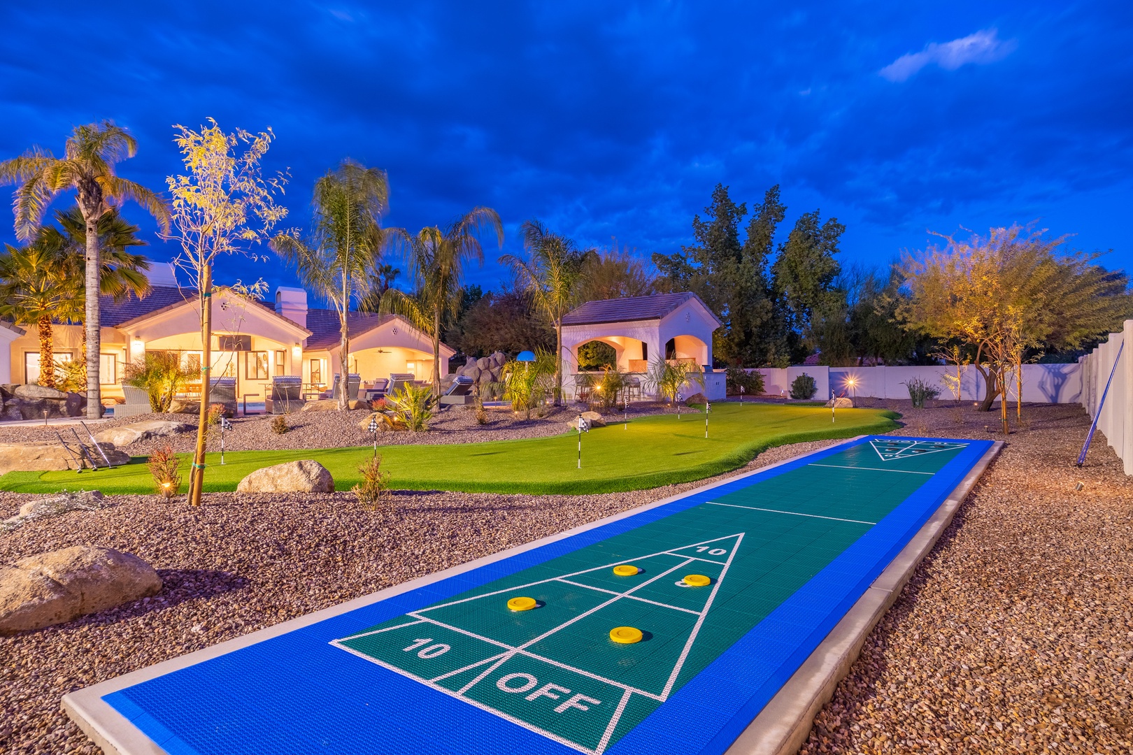 Expansive estate with shuffleboard court, putting green, lush landscaping, and glowing evening ambiance under vibrant twilight skies.